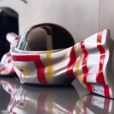 Escultura de un casco envuelto en tela blanca con franjas rojas y amarillas, firmada Polito